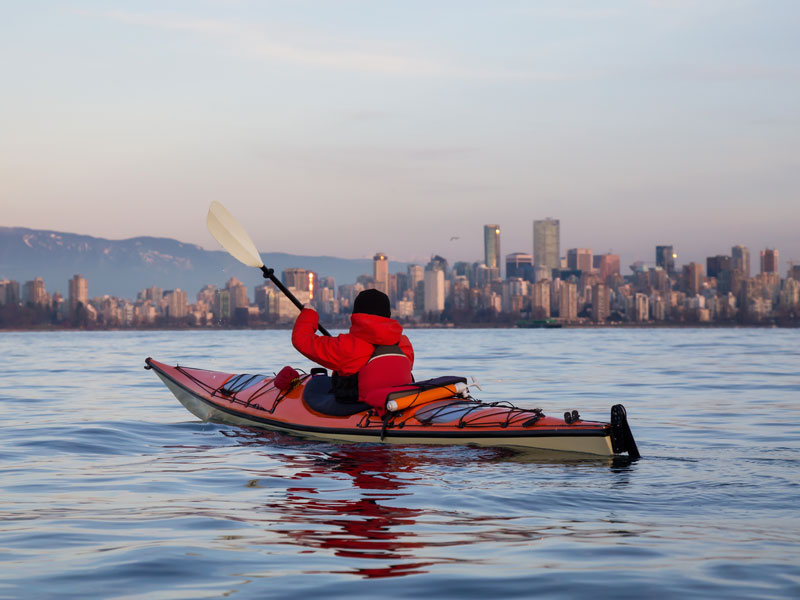 A man paddling a sea-kayak in winter with a city in the background.