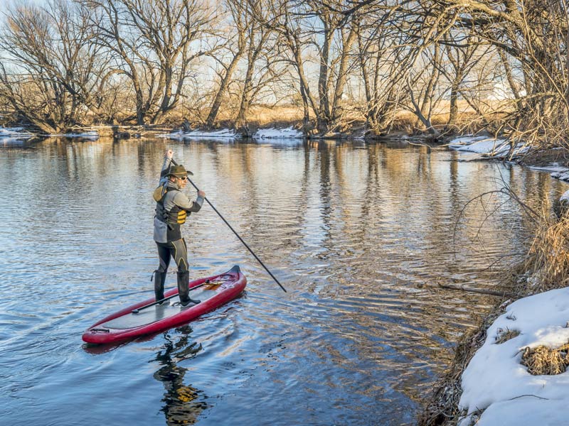 A man wearing a dry suit and warm clothes paddling a SUP on an icy river in winter