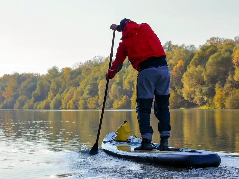 A person paddle boarding down a autumnal river scene wearing layers of clothing