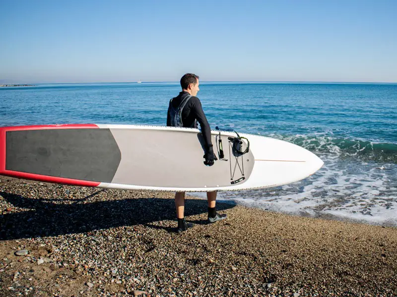 A man carrying one of the types of paddle boards into the sea, a touring paddle board.